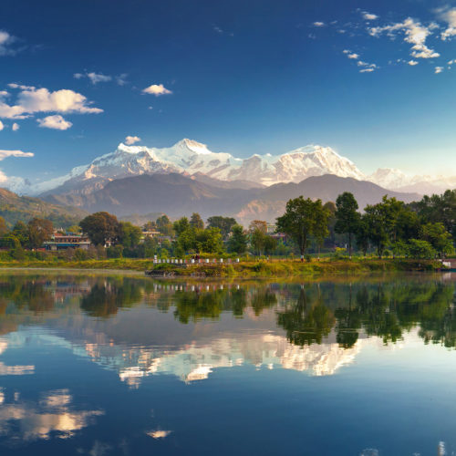 Scenic view of Phewa Lake in Pokhara with the reflection of the surrounding hills and snow-capped mountains in the calm water.