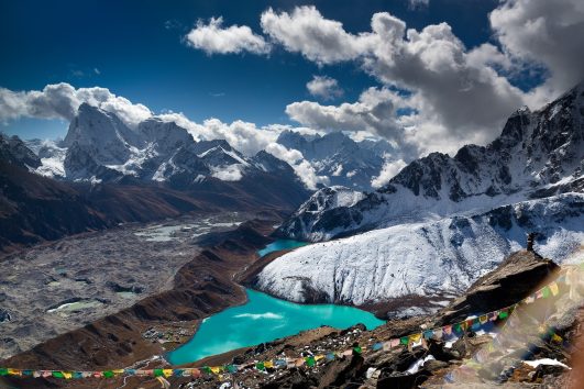 A breathtaking view of Gokyo Lake, a serene turquoise glacial lake surrounded by towering snow-capped peaks in the Everest region of Nepal. The clear blue sky reflects on the calm waters, with rugged mountain landscapes and rocky terrain adding to the dramatic beauty of the scene.