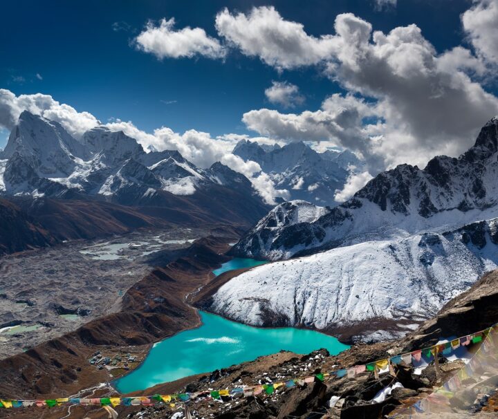 A breathtaking view of Gokyo Lake, a serene turquoise glacial lake surrounded by towering snow-capped peaks in the Everest region of Nepal. The clear blue sky reflects on the calm waters, with rugged mountain landscapes and rocky terrain adding to the dramatic beauty of the scene.